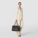 Front view: A woman carries the dark brown, geometrically woven SECRET TOTE BAG IN RIBBON MOSAICO by its handles; “SERAPIAN” is written on the bag.