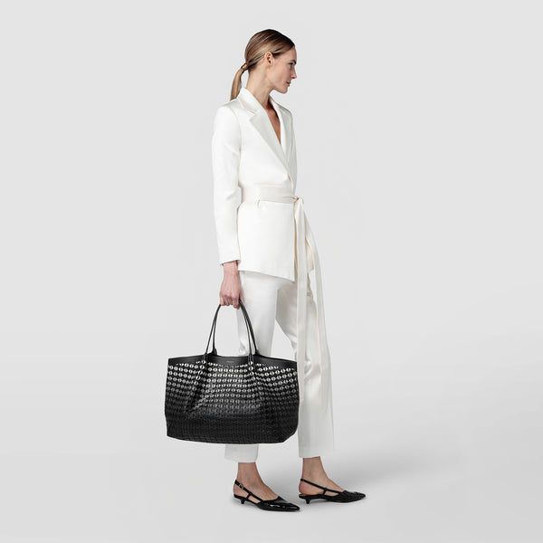 Side view of a woman in a white suit carrying the SECRET TOTE BAG IN MOSAICO made of black-patterned, handwoven nappa leather on her arm.
