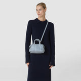 Front view: A woman wears the MINI ANI BAG IN RIBBON MOSAICO, with its light blue three-dimensional woven pattern and short handles, across her shoulder with a navy dress.