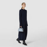 Front view: A woman carries the MINI ANI BAG IN RIBBON MOSAICO in Arctic Blue by its handle; the small bag features a dimensional geometric weave and gold-tone details.