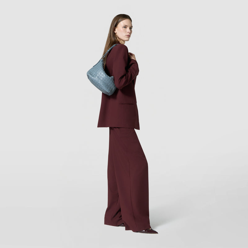 Side view: A woman carries the SMALL ANI SHOULDER BAG IN MOSAICO in Storm Blue on her shoulder, paired with a burgundy pantsuit.