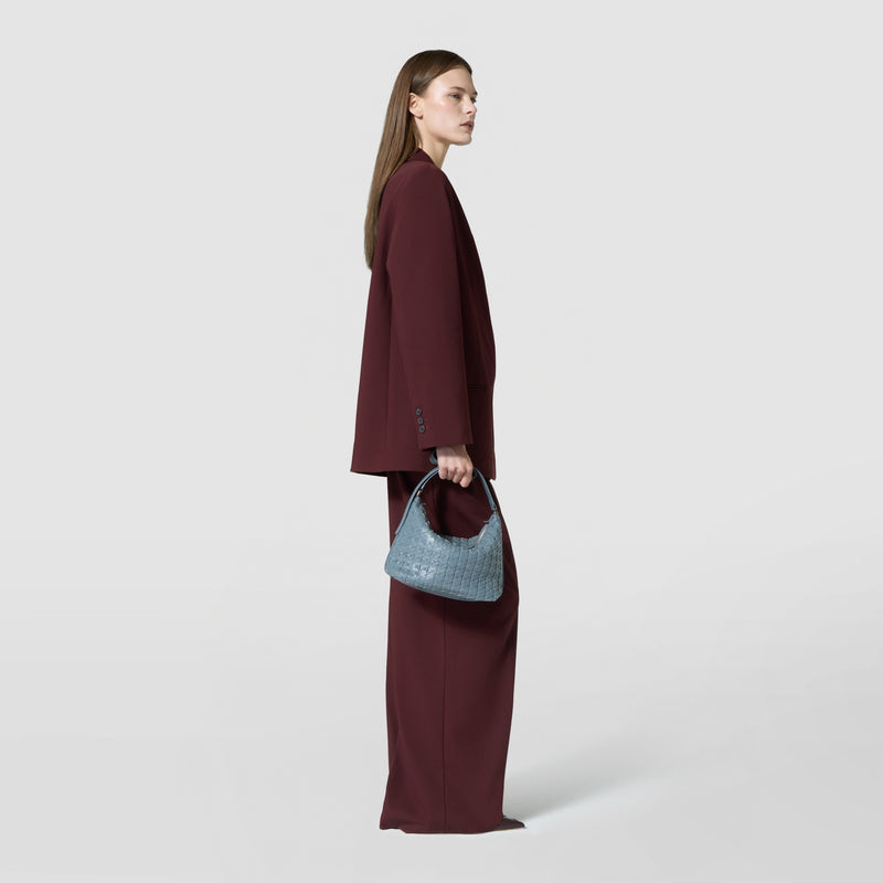 Side view of a person in a burgundy suit holding the SMALL ANI SHOULDER BAG IN MOSAICO in Storm Blue in their left hand.