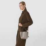 Front view: A woman carries the MINI BUCKET IN MOSAICO in light taupe leather with a distinctive woven pattern, short handles, and a shoulder strap, paired with a dark brown outfit.