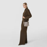 Front view: A woman carries the MINI BUCKET IN MOSAICO, made of taupe geometric-weave leather with short handles and a long shoulder strap, with a dark brown outfit.