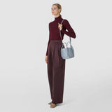 Front view: A woman wears the MINI BUCKET IN RIBBON MOSAICO in ice blue, featuring a three-dimensional woven texture and a long shoulder strap, at her side.
