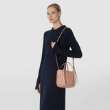 Front view: A woman carries the MINI BUCKET IN MOSAICO in sandal-colored leather with a geometric woven pattern and a narrow shoulder strap, paired with a dark blue dress.