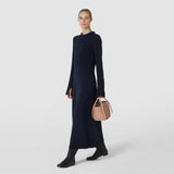 Front view: A woman carries the MINI BUCKET IN MOSAICO in sandalwood-colored, intricately woven leather by its short handles; the soft bucket bag hangs beside her dark dress.