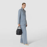 Front view: A woman wears the BUCKET IN MOSAICO bag in black woven leather with narrow straps, paired with a light blue pantsuit against a light background.