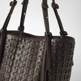 Dettaglio: BUCKET IN MOSAICO RIBBON mostra la sua superficie in pelle scura finemente intrecciata e i sottili manici a spalla su uno sfondo chiaro.