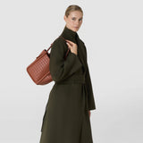 Front view: A woman wears the BUCKET IN MOSAICO in brick-red woven leather over her shoulder; the bag features a soft shape and narrow straps.