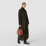 BUCKET IN MOSAICO: Front view of a woman carrying a brick-red bucket bag with an embossed mosaic pattern and knotted straps beside a dark coat.