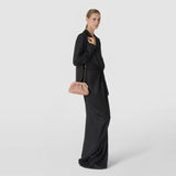 Front view: MINI SECRET CLUTCH BAG IN MOSAICO hangs from a gold-tone chain beside a woman. The sand-colored clutch with a raised mosaic pattern complements her dark pinstripe outfit in a studio.