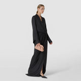 The MINI SECRET CLUTCH BAG IN MOSAICO, a small sandalwood-colored clutch with a geometric woven pattern, is held by an elegantly dressed woman; front view against a light background.