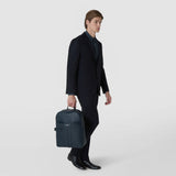 BACKPACK IN STEPAN in ocean blue, with an embossed pattern, front pocket and side strap; front view, held by the top handle by an elegantly dressed man.
