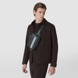 Front view: A model wears the SLING BACKPACK IN STEPAN across the chest. The asphalt-gray backpack features an S-etching, ebony and ice-blue stripes, and “SERAPIAN”.