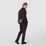 Side view: A man wears the CAMERA CROSSBODY BAG IN STEPAN across his shoulder; the dark gray, textured bag with a light blue center stripe stands out against a neutral studio background.