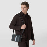 Frontal view: A man wears the CAMERA CROSSBODY BAG IN STEPAN over his shoulder. The dark gray bag features an S pattern, leather details, and light blue stripes. Inscription: “SERAPIAN”.