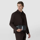 Front view: A man holds the WASHBAG IN STEPAN in front of his dark brown coat. The black embossed bag has a light blue leather strip. Inscription: SERAPIAN.