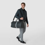 Front view: A man carries the HOLDALL IN STEPAN by its handles and over his shoulder; the asphalt-gray bag features a textured surface and brown and ice-blue stripes.