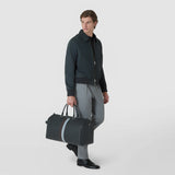 HOLDALL IN STEPAN in asphalt gray with an embossed pattern and contrasting stripes is carried by a man by the double handles; front view against a light studio background.