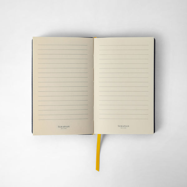 Front view of opened NOTEBOOK IN CACHEMIRE LEATHER with lined pages, yellow bookmark, and white paper. &#39;SERAPIAN MILANO&#39; printed at the bottom.